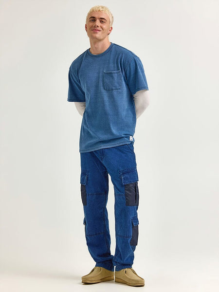 A person stands wearing the Alpha x Lee Looser Fit Indigo Dyed Tee by Alpha Industries over a long-sleeve shirt, complemented by blue cargo pants featuring reinforced pockets and patches, along with tan shoes.