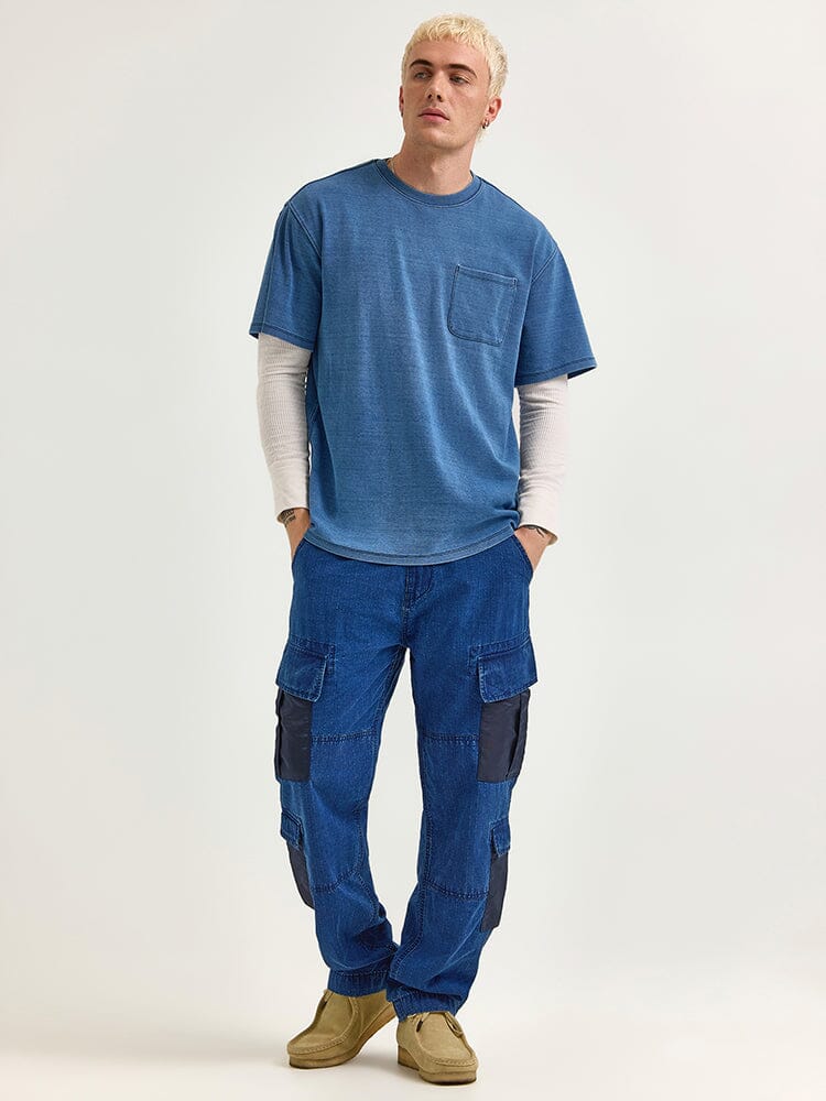A person wearing an Alpha Industries FW24-Lee Looser Fit Indigo Dyed Tee over a white long-sleeve shirt, paired with blue cargo pants featuring reinforced pockets, and tan shoes.