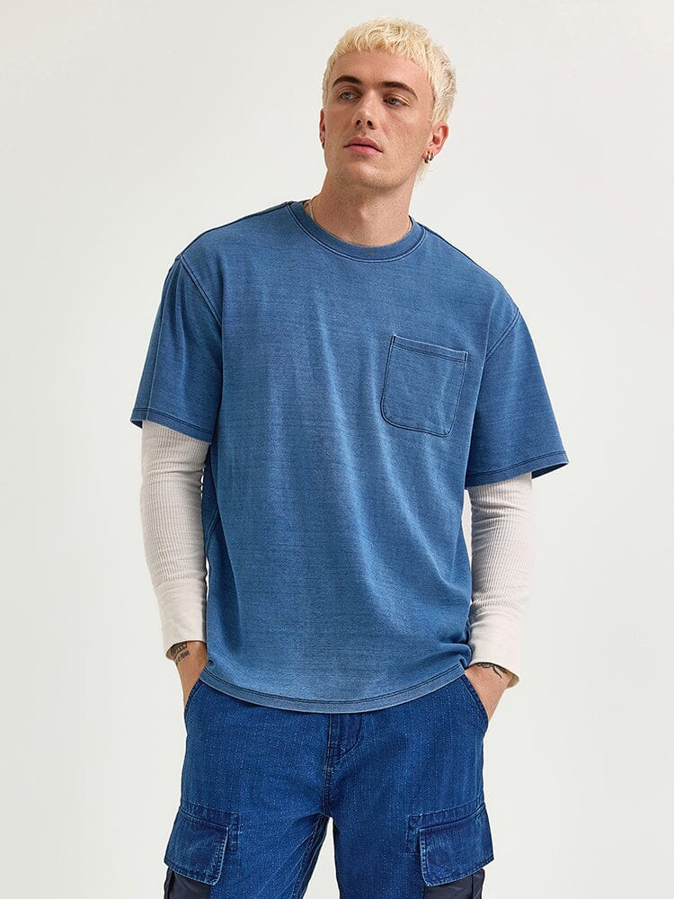 A person wearing an Alpha Industries' Fw24-Lee Looser Fit Indigo Dyed Tee with reinforced pocket details over a white long-sleeve shirt and blue cargo pants.