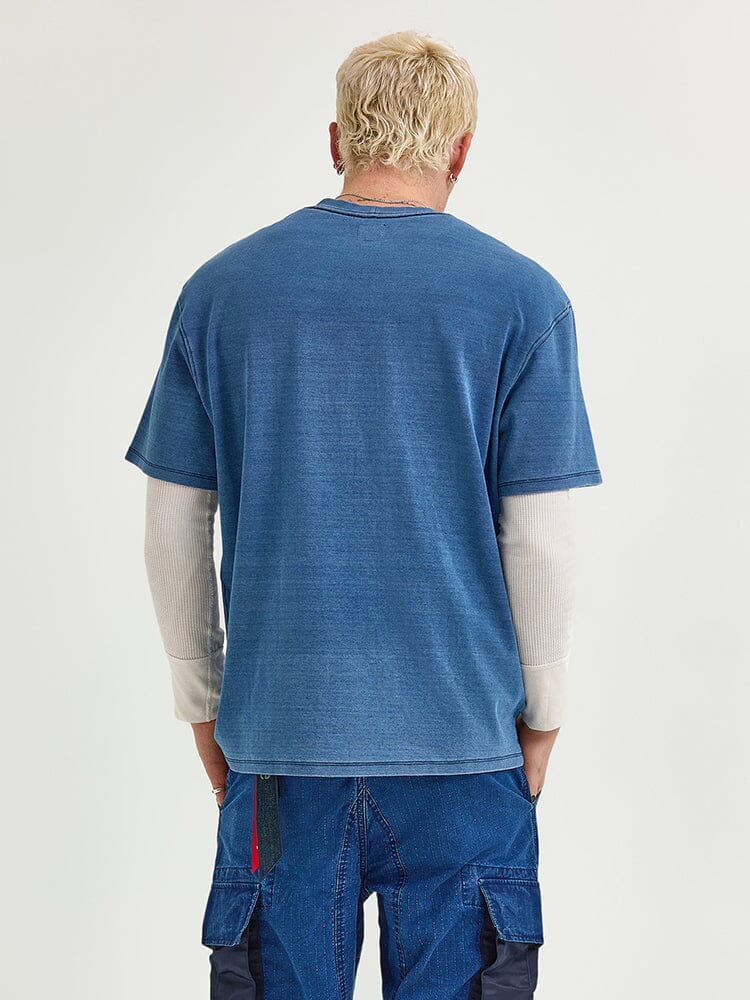 A person with short blond hair is seen from behind wearing an Alpha Industries FW24-Lee Looser Fit Indigo Dyed Tee over a white long-sleeve shirt and blue pants.