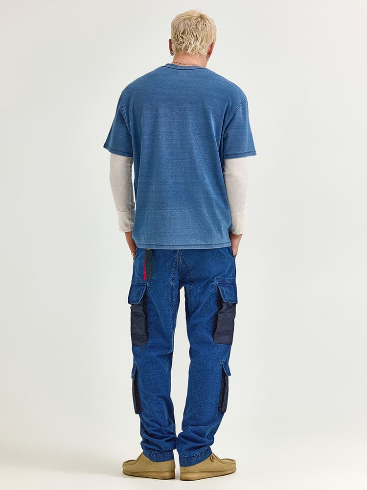 A man stands facing away, wearing the Alpha Industries Fw24-Lee Looser Fit Indigo Dyed Tee layered over a long-sleeve shirt, paired with blue cargo pants featuring reinforced pockets and black patches, complemented by light brown shoes.