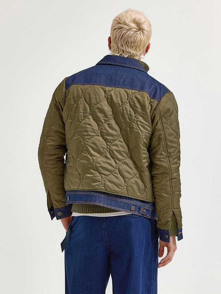 A person wearing the Alpha X Lee Quilted Jacket by Alpha Industries is exhibiting a heritage style with green quilted fabric and denim accents, complemented by blue pants. They stand with their back to the camera, capturing a mixed-media approach to fashion.