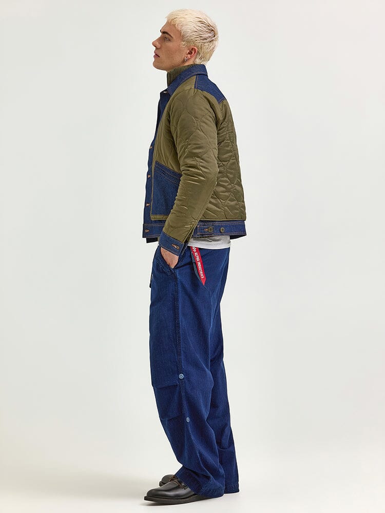 In profile, a person with blond hair is wearing an Alpha X LEE Quilted Jacket from Alpha Industries, exuding a heritage style. The outfit is completed with blue wide-leg jeans and black shoes.