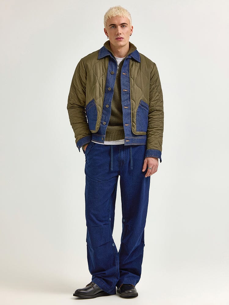 A person stands showcasing a heritage style with an Alpha X Lee Quilted Jacket by Alpha Industries in green quilted fabric and blue trim, paired with a gray sweater, blue pants, and black shoes.