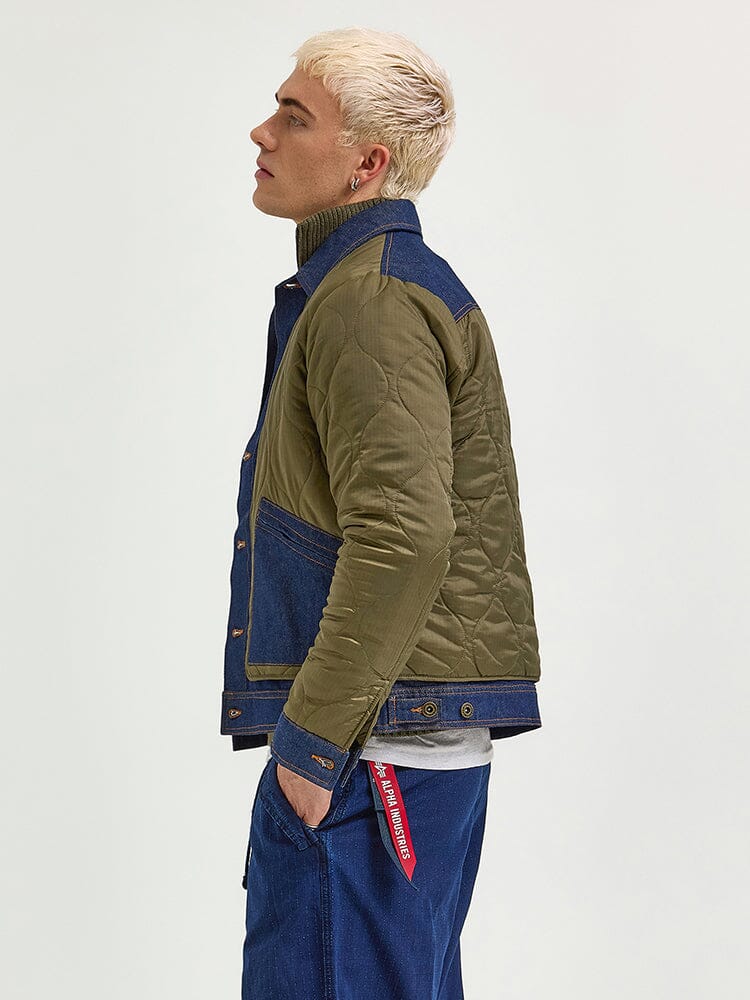 An individual with short blonde hair is wearing an Alpha Industries quilted jacket, blending green and denim, paired with blue jeans and a red tag accessory. They stand highlighting a mixed-media approach with heritage style.