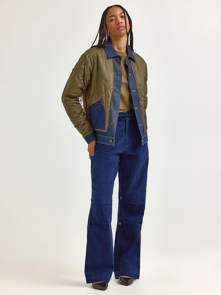 Clad in the heritage-themed Alpha X Lee Quilted Jacket from Alpha Industries, the individual exudes confidence. The jacket boasts a green quilted design accented with blue details, perfectly complemented by blue jeans and black shoes.