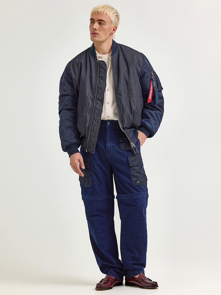 A person with short blond hair is wearing the Alpha X Lee Reversible MA-1 Jacket by Alpha Industries in blue, along with a white shirt and blue cargo pants.