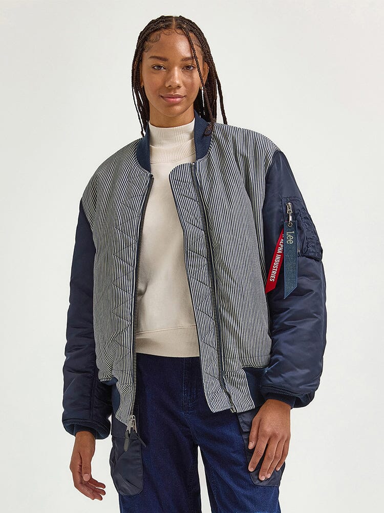 A person with braided hair is wearing the Alpha X Lee Reversible MA-1 Jacket from Alpha Industries, styled over a white turtleneck and blue jeans.