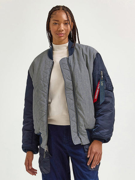 A person with braided hair is wearing the Alpha X Lee Reversible MA-1 Jacket from Alpha Industries, styled over a white turtleneck and blue jeans.
