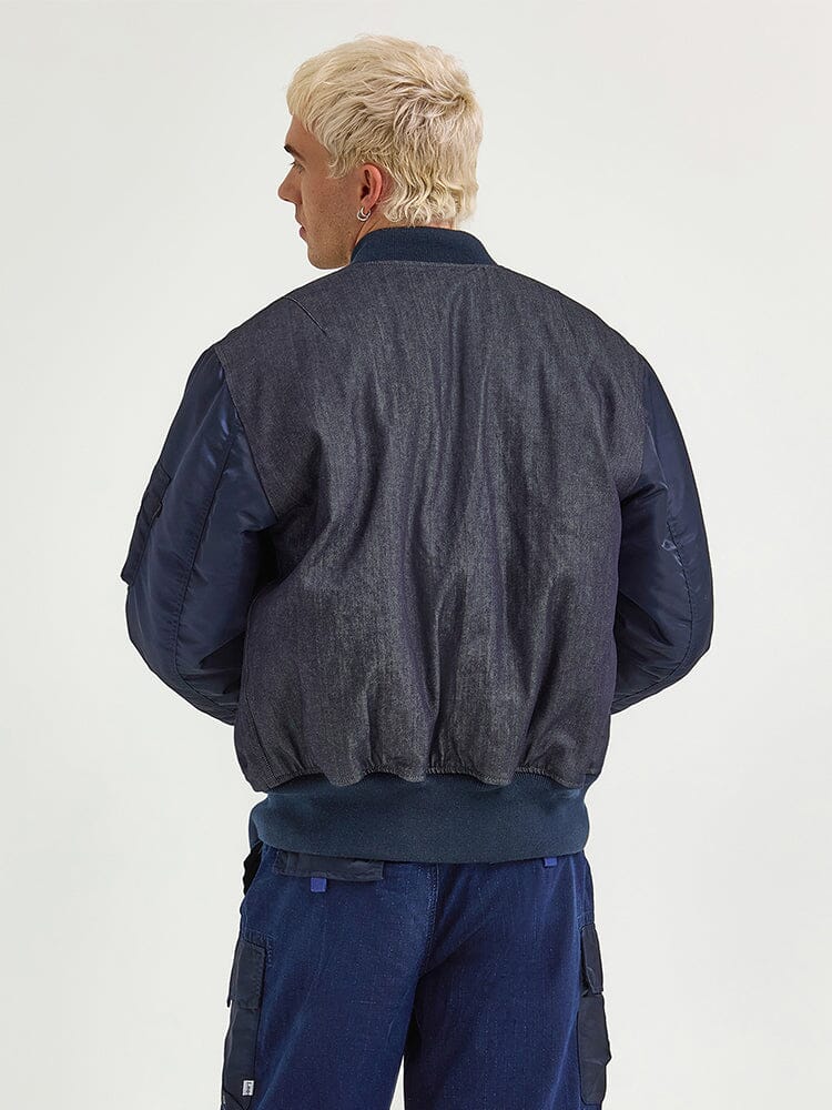 A person with light hair, seen from the back, is wearing a dark vintage railroad-stripe bomber jacket with blue sleeves and matching pants, specifically the Alpha X Lee Reversible MA-1 Jacket by Alpha Industries.