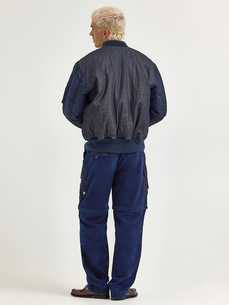 A person with short blond hair is wearing the Alpha X Lee Reversible MA-1 Jacket by Alpha Industries and blue cargo pants, standing with their back to the camera.