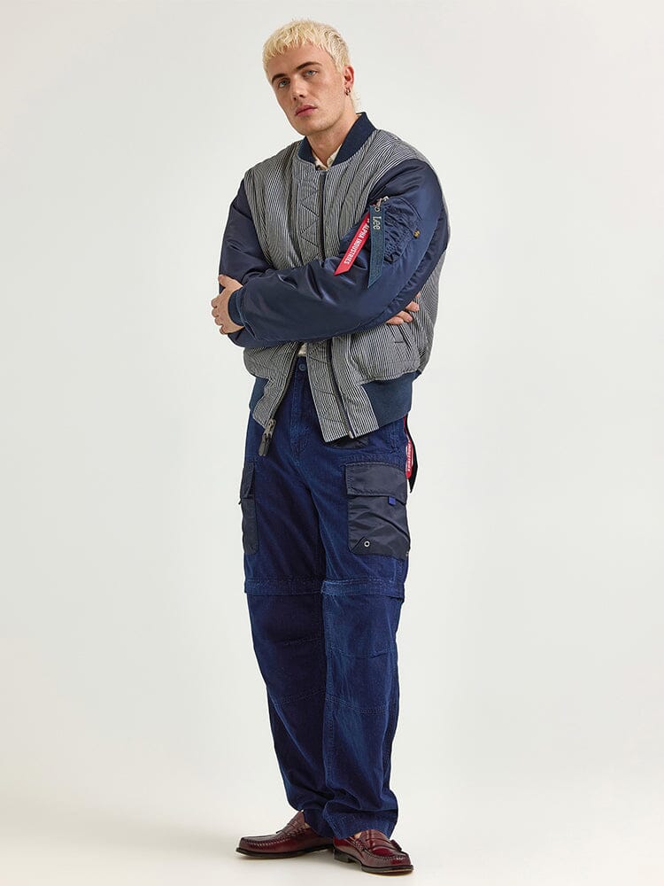 A person stands confidently with arms crossed, wearing an Alpha X Lee Reversible MA-1 Jacket by Alpha Industries, matched with blue cargo pants and maroon shoes.