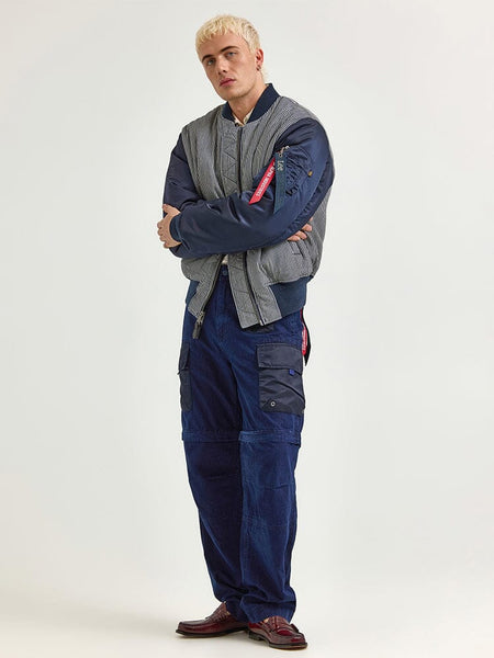 A person stands confidently with arms crossed, wearing an Alpha X Lee Reversible MA-1 Jacket by Alpha Industries, matched with blue cargo pants and maroon shoes.