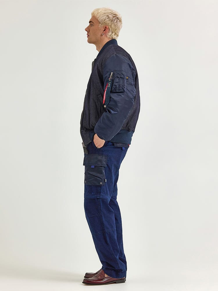 A person with short, light hair stands in profile wearing the stylish Alpha X Lee Reversible MA-1 Jacket by Alpha Industries, along with blue cargo pants and brown shoes.