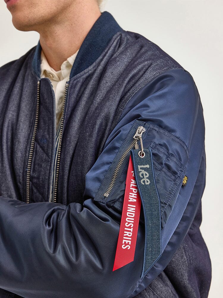 A person wears the Alpha X Lee Reversible MA-1 Jacket by Alpha Industries, featuring a navy design with a red Alpha Industries tag hanging from the sleeve zipper, highlighting its versatile style similar to that of a reversible jacket.