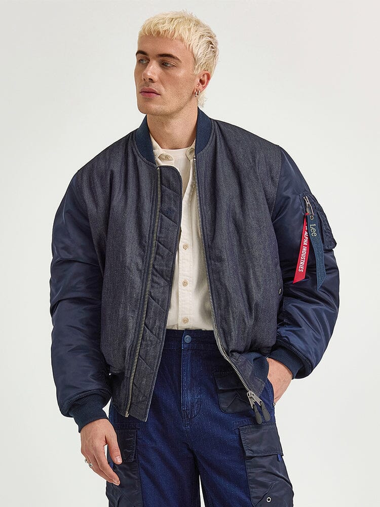 The individual sports an Alpha X Lee Reversible MA-1 Jacket by Alpha Industries over a light shirt and blue pants, gazing thoughtfully to the side.