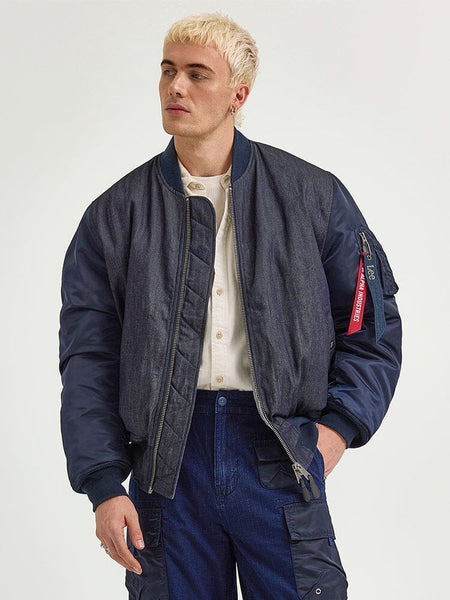 The individual sports an Alpha X Lee Reversible MA-1 Jacket by Alpha Industries over a light shirt and blue pants, gazing thoughtfully to the side.