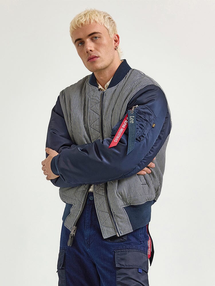A person with blond hair wearing an Alpha Industries FW24-Lee Alpha X Lee Reversible MA-1 Jacket and blue pants stands with arms crossed.