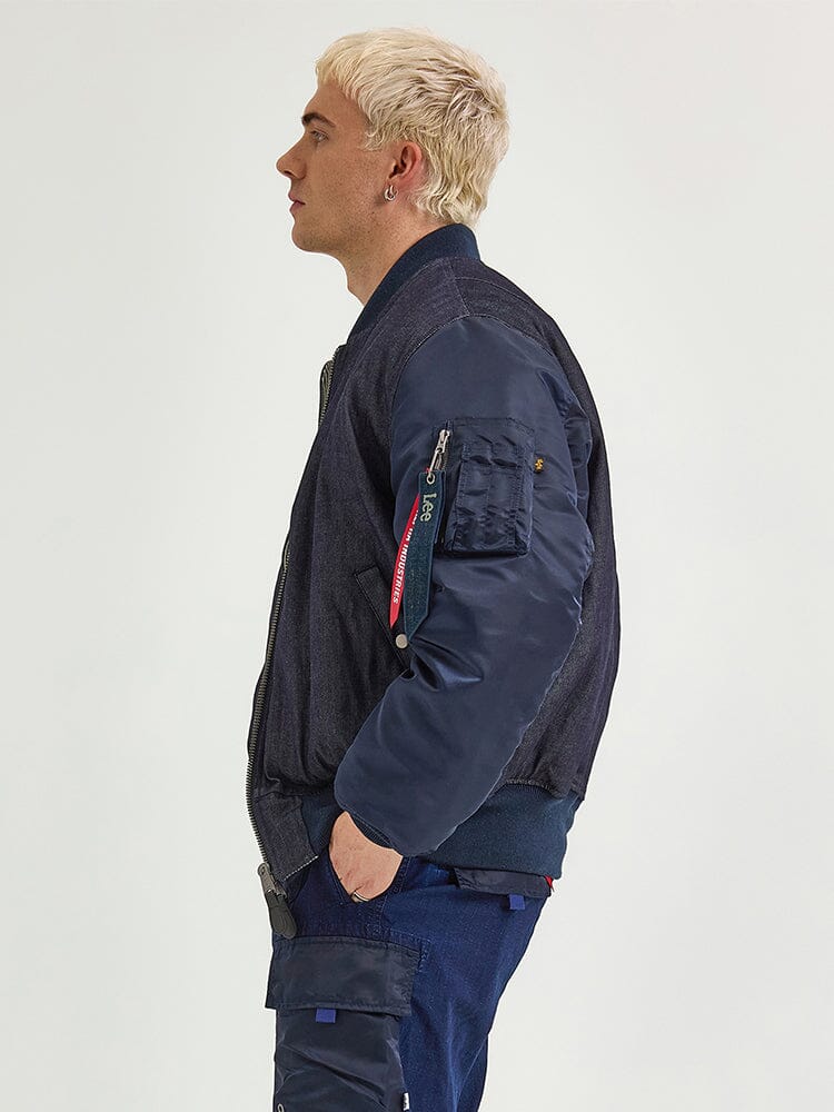 A person with short blond hair stands in profile, wearing the Alpha X Lee Reversible MA-1 Jacket by Alpha Industries, along with blue cargo pants.