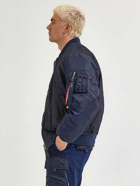 A person with short blond hair stands in profile, wearing the Alpha X Lee Reversible MA-1 Jacket by Alpha Industries, along with blue cargo pants.