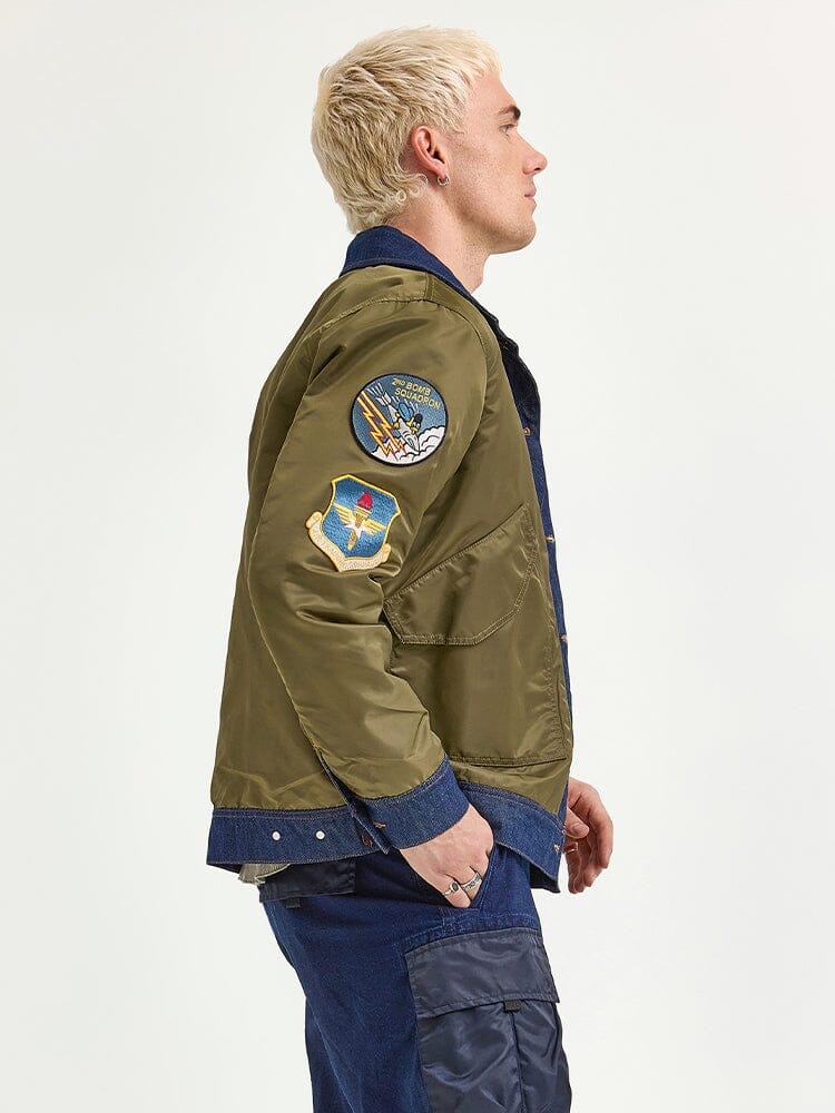 A person with short blond hair is seen in a side profile view wearing the olive green Alpha X Lee Reversible Rider Jacket by Alpha Industries, featuring patches on the sleeve, paired with dark pants.