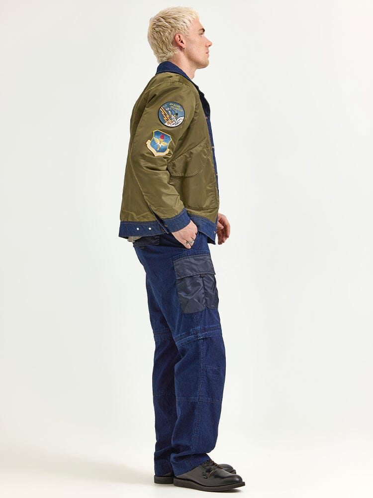 A person with blonde hair stands sideways, wearing the Alpha X Lee Reversible Rider Jacket by Alpha Industries in green, along with patches, blue cargo pants, and black shoes.