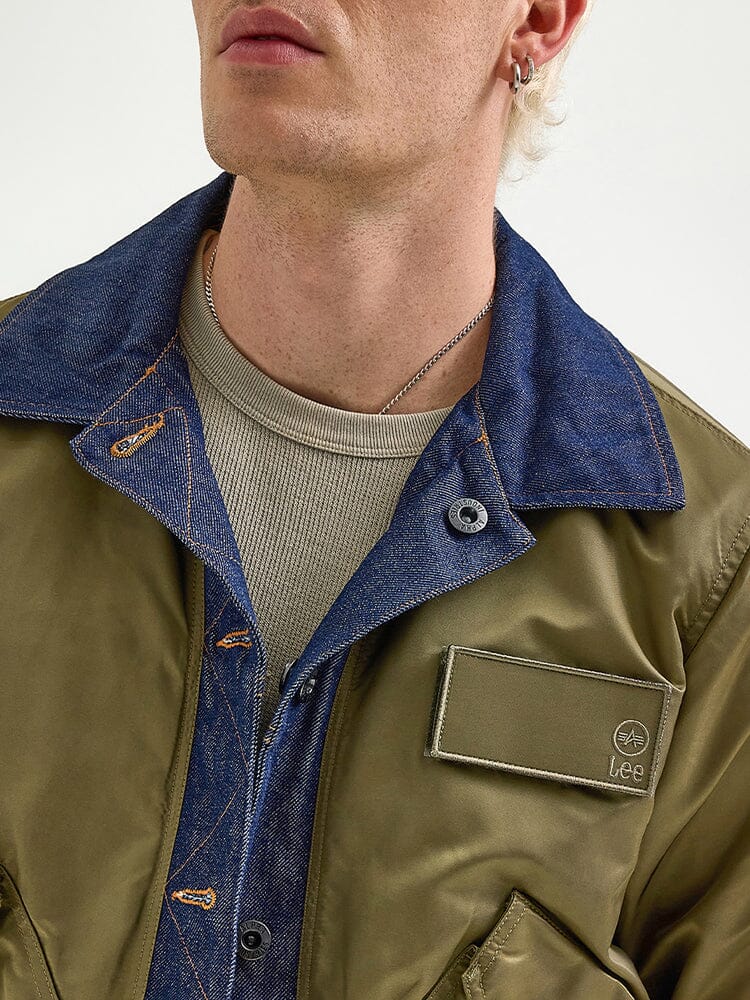 An individual wearing an olive green Alpha X Lee Reversible Rider Jacket by Alpha Industries, featuring a denim lining, adorned with a button and badge on the chest, paired with a beige shirt underneath and accessorized with a chain necklace and earring.
