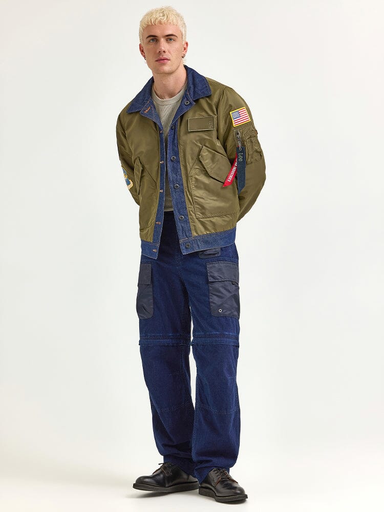 A person with short blonde hair is wearing the Alpha X Lee Reversible Rider Jacket by Alpha Industries, featuring green and blue sides with patches. They complement this with blue pants that have pockets and black shoes.