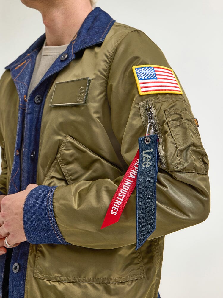 A person wearing an Alpha Industries x Lee reversible Rider Jacket, featuring a U.S. flag patch on the sleeve and a red Alpha Industries tag, layered over a denim jacket.