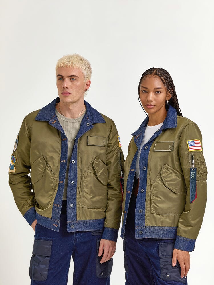 Against a plain backdrop, two models stand side by side wearing matching indigo Alpha x Lee Reversible Rider Jackets from Alpha Industries, each adorned with patches.