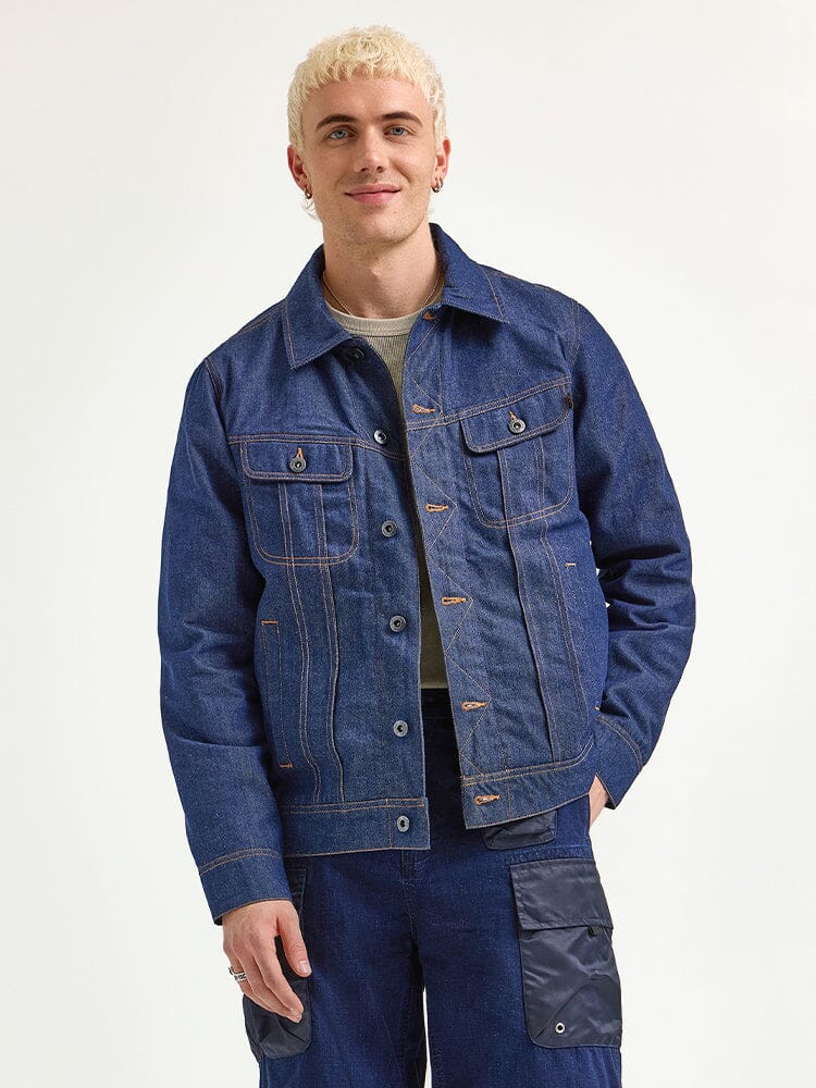 A person with short blond curly hair is wearing an Alpha Industries x Lee Reversible Rider Jacket and denim pants.