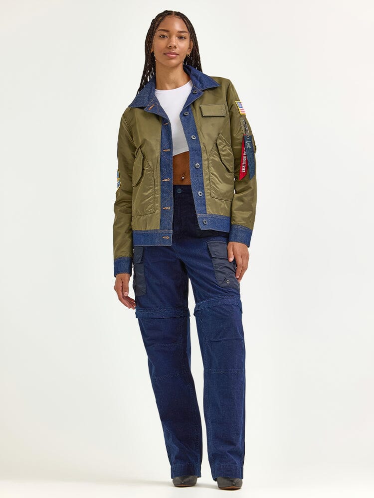A person stands wearing an Alpha X Lee Reversible Rider Jacket paired with a white crop top and blue cargo pants.
