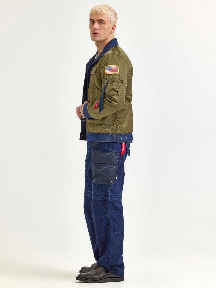 A person with short blonde hair is wearing an olive green Alpha Industries Alpha x Lee Reversible Rider Jacket featuring an American flag patch, along with blue cargo pants.