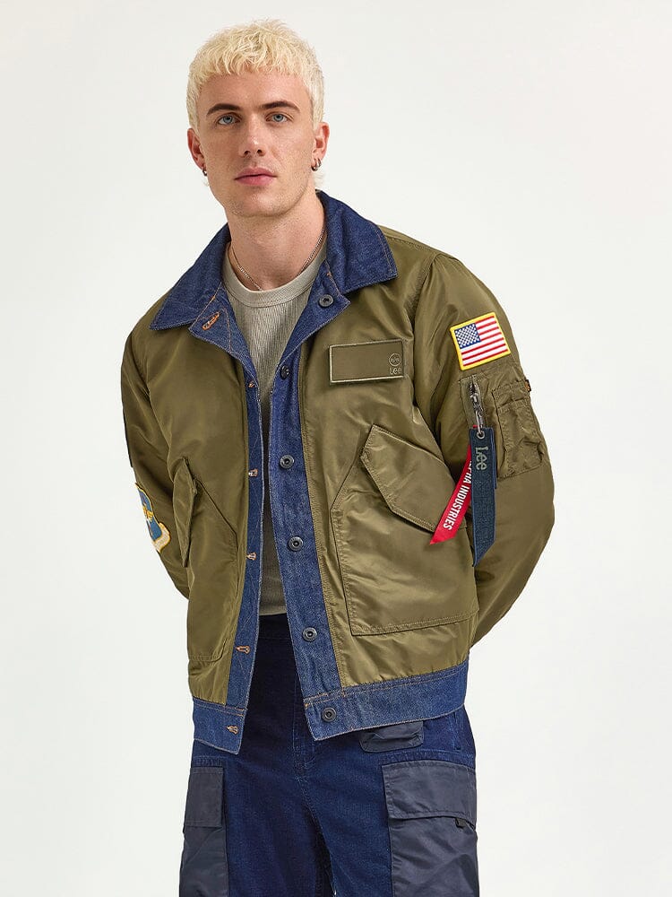 A person with short blond hair wears the Alpha X Lee Reversible Rider Jacket by Alpha Industries, featuring green and denim fabrics adorned with patches such as an American flag, paired with dark pants.