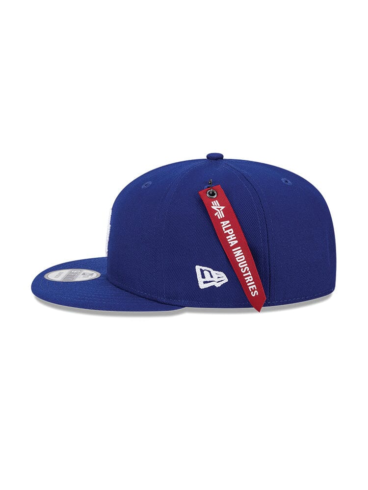 The Los Angeles Dodgers X Alpha X New Era 950 Cap (Blue) from Alpha Industries under model number FW24-NE-MLB boasts a stylish curved brim and prominently displays a red tag at the back.