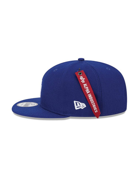 The Los Angeles Dodgers X Alpha X New Era 950 Cap (Blue) from Alpha Industries under model number FW24-NE-MLB boasts a stylish curved brim and prominently displays a red tag at the back.