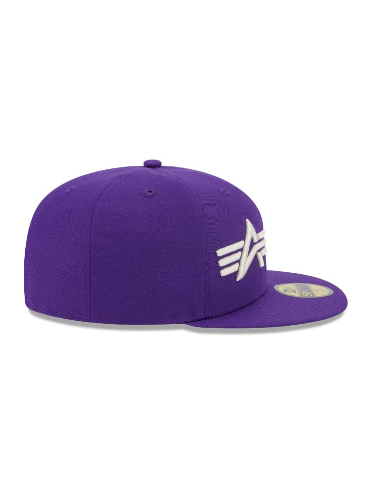 A Los Angeles Laker x Alpha x New Era 59FIFTY Fitted Cap by Alpha Industries, featuring a purple base with a white embroidered design on the side, is resting on a grey surface.