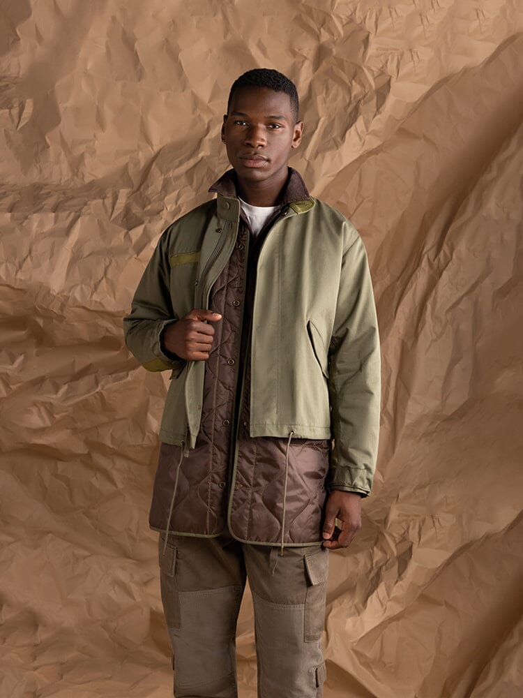 A person wearing the M-65 Long Liner by Alpha Industries. The jacket features an onion quilted lining over a brown quilted coat paired with gray cargo pants, highlighting its 3-in-1 layering versatility.
