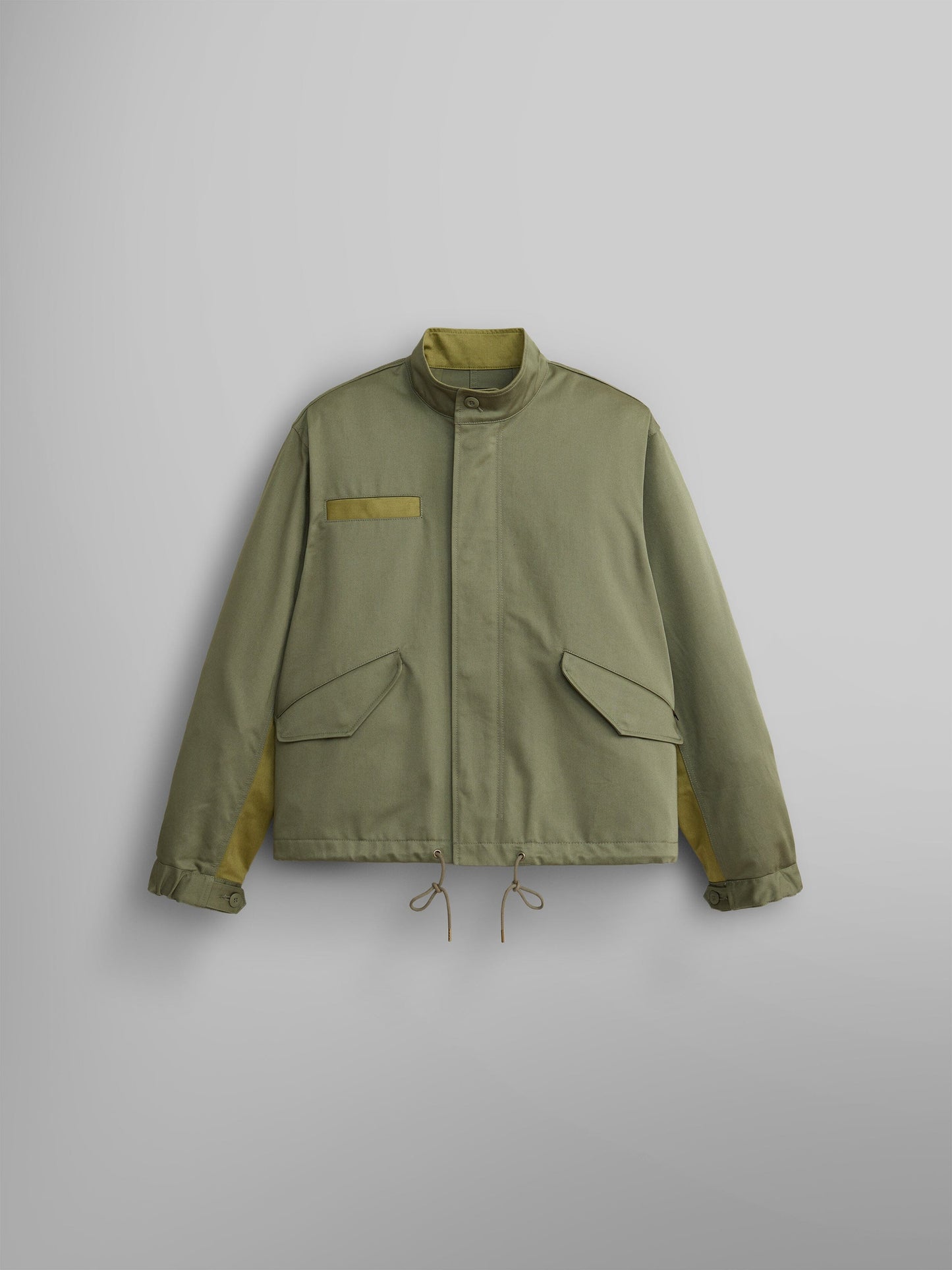 The M-65 Long Liner by Alpha Industries is a light green jacket made from durable NYCO fabric. It features a high collar, buttoned front, adjustable cuffs, and a drawstring hem.