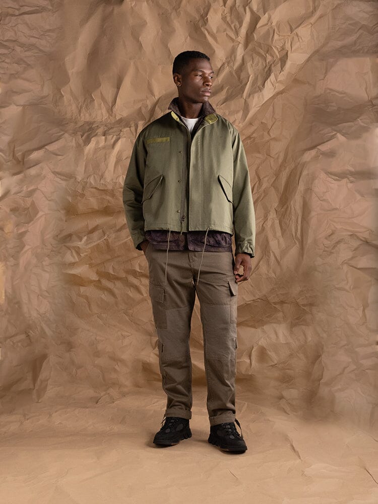 An person wearing the M-65 Long Liner in green, featuring an onion quilted lining from Alpha Industries, paired with brown pants.