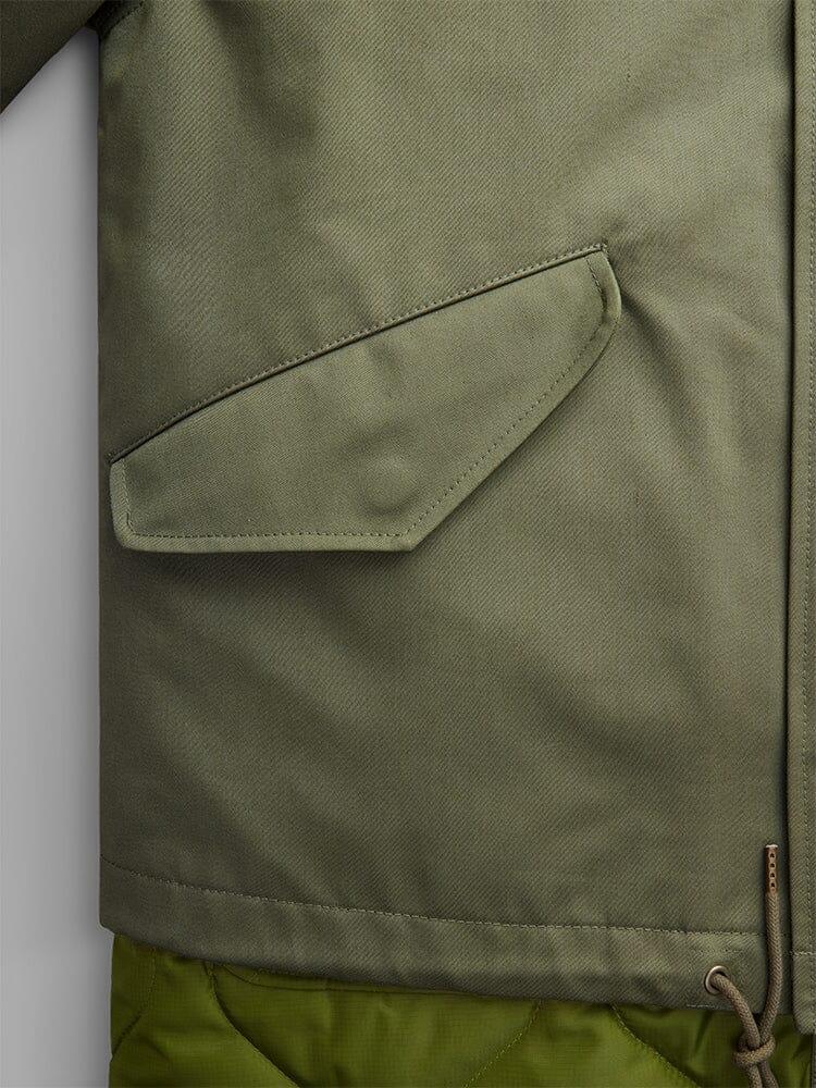 Close-up of the M-65 Long Liner in OG-107 Green by Alpha Industries showcasing its precise stitching, a side flap pocket, and a bottom drawstring with a metal tip.