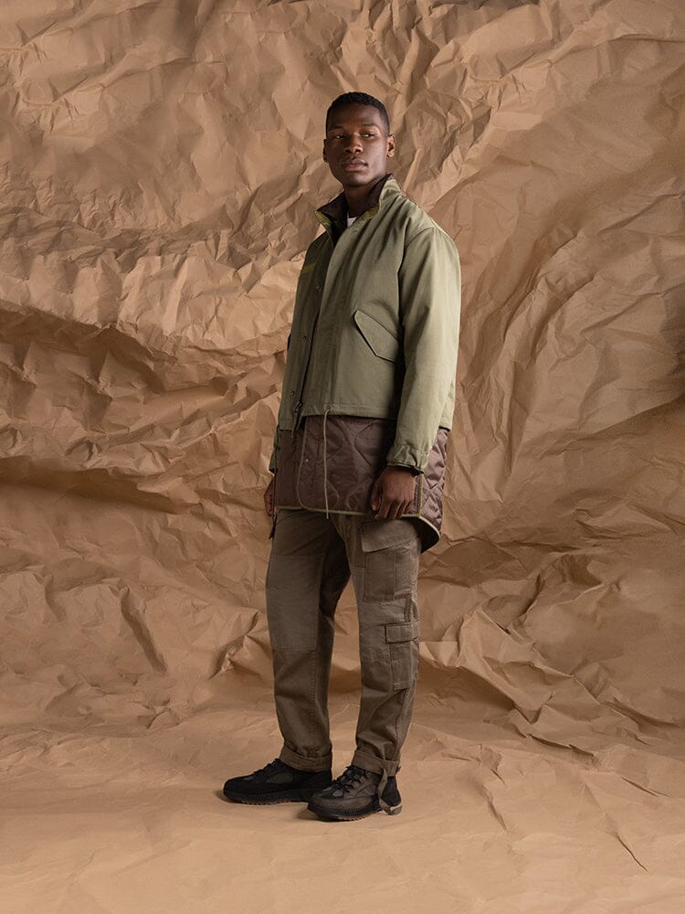 A person stands wearing the M-65 Long Liner in olive green from Alpha Industries, which features onion quilted lining.
