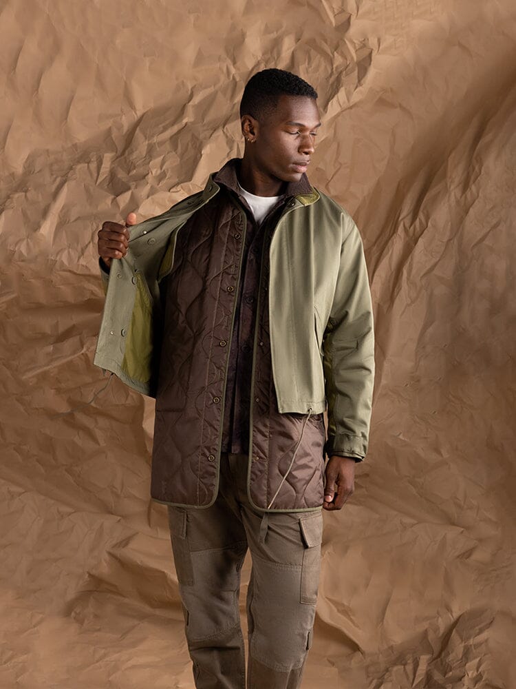 A person wearing the M-65 Long Liner jacket by Alpha Industries, showcasing its 3-in-1 layering over a brown quilted vest, looking to the side.