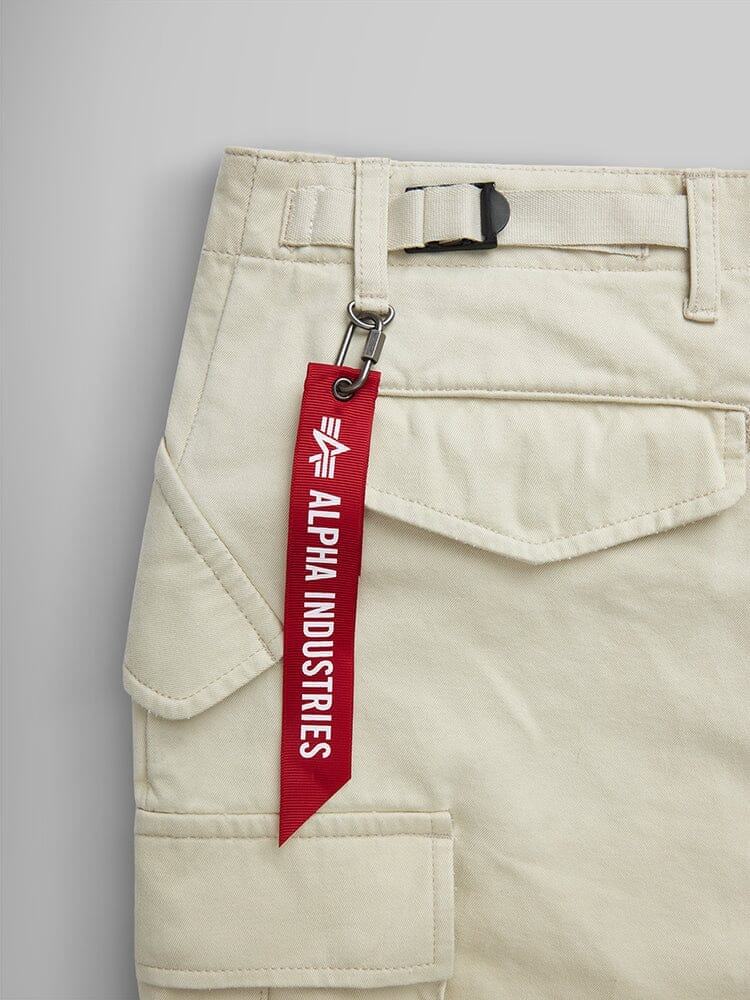 Alpha Industries M-65 Pant in beige with utility pockets, black buckle belt, and red Alpha Industries keychain near the waistband.