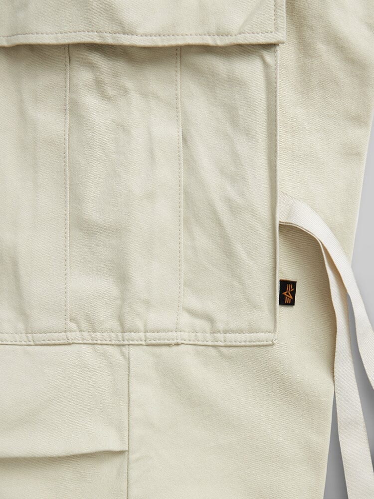 Alpha Industries M-65 Pant in beige canvas with utility pockets, detailed stitching, waist strap, and small black tag with orange design.