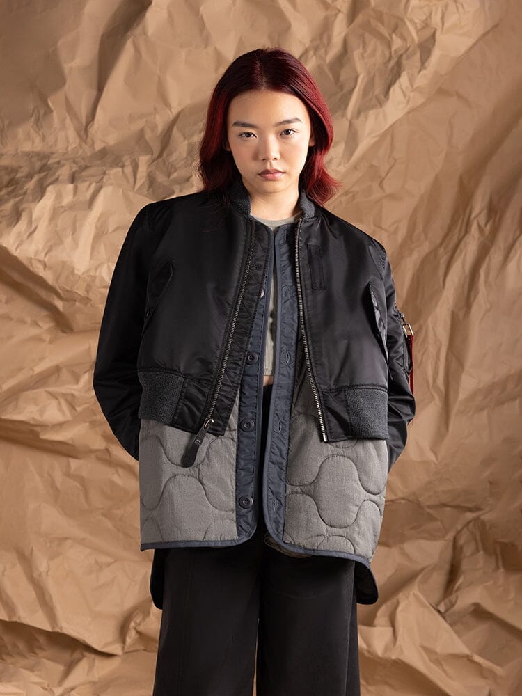 A person stands wearing the Women's 3-in-1 MA-1 Quilted Liner Jacket in black and gray by Alpha Industries.