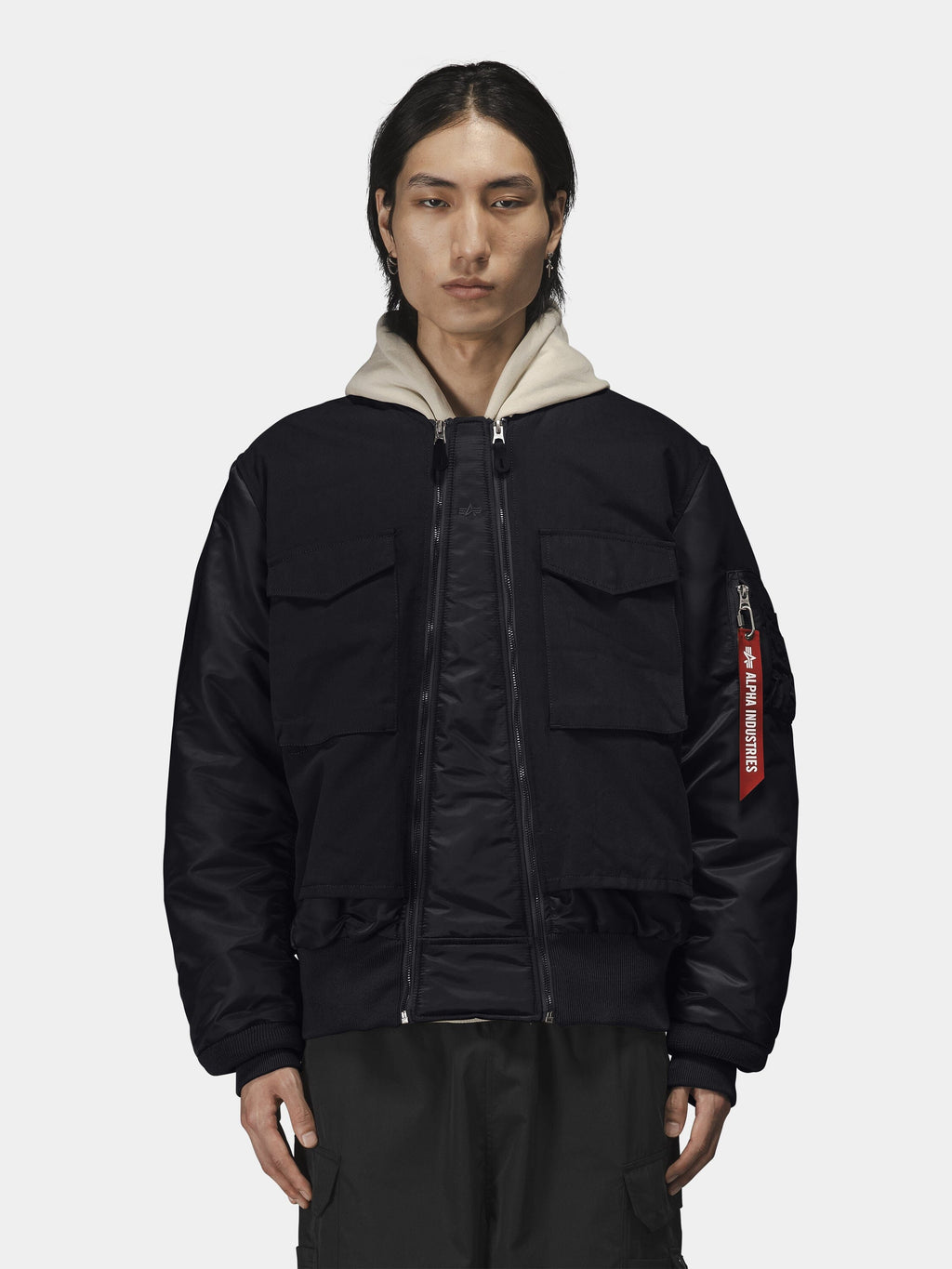 Alpha Industries MA-1 Mixed Cotton Flight Jacket in black with large front pockets and a red sleeve tag, over a beige hoodie.