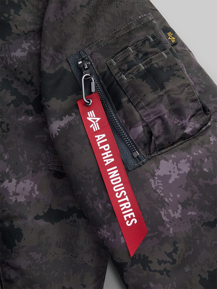 Alpha Industries MA-1 Mod Bomber Jacket sleeve with camouflage print, zip utility pocket, and red Alpha Industries tag on zipper pull.