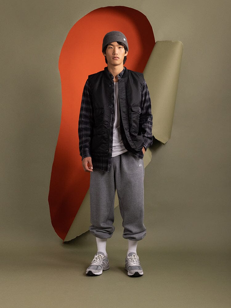 A person stands wearing a beanie, the Alpha Industries MA-1 Mod Vest in black with quilted lining, a plaid shirt, gray sweatpants.
