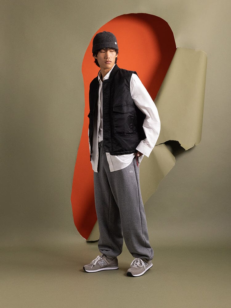 A person stands wearing the black MA-1 Mod Vest from Alpha Industries with a beanie, white shirt, and gray sweatpants.